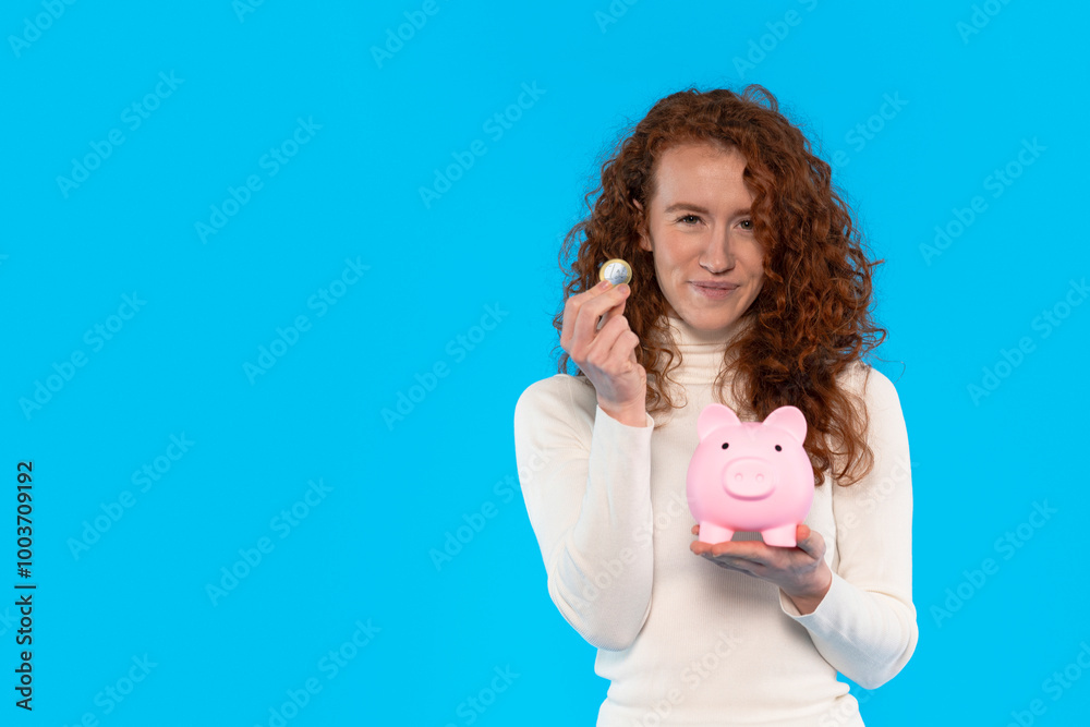 A young woman joyfully saving coins in a playful pink piggy bank against a bright blue backdrop A young woman joyfully saving coins in a playful pink piggy bank against a bright blue backdrop