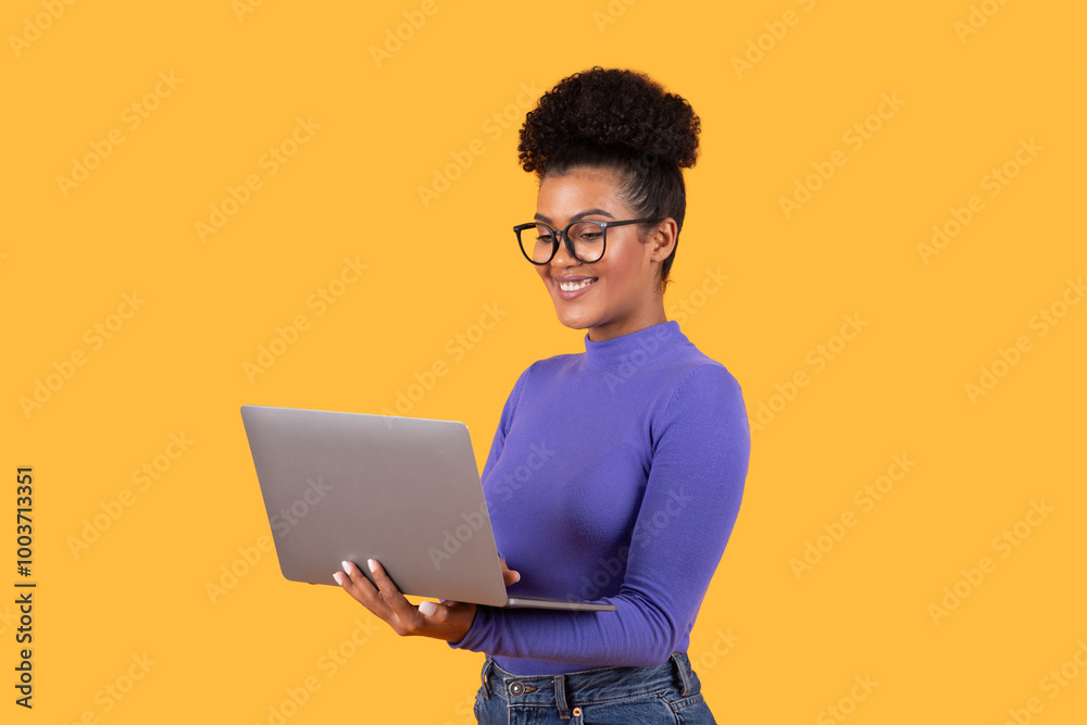 A young woman with curly hair and glasses smiles while using her laptop, standing in front of a vivid yellow background. Her cheerful demeanor suggests a productive moment in her work. A young woman with curly hair and glasses smiles while using her laptop, standing in front of a vivid yellow background. Her cheerful demeanor suggests a productive moment in her work.