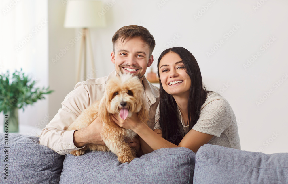 Portrait of happy laughing young couple close near couch with cute little dog, smiling husband and wife with puppy, relaxing together in new own living room, apartment renters with companion animal Portrait of happy laughing young couple close near couch with cute little dog, smiling husband and wife with puppy, relaxing together in new own living room, apartment renters with companion animal