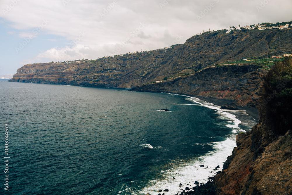 Black sandy and volcanic rocky coast with blue color of water. Shore of Atlantic Ocean in Puerto de la Cruz. Travel concept. Amazing view of Tenerife island north coast. Popular tourist destination. Black sandy and volcanic rocky coast with blue color of water. Shore of Atlantic Ocean in Puerto de la Cruz. Travel concept. Amazing view of Tenerife island north coast. Popular tourist destination.