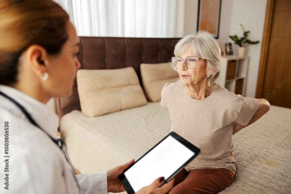Senior woman with backache sitting on bed at home while her doctor is listening symptoms Senior woman with backache sitting on bed at home while her doctor is listening symptoms