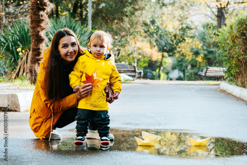 Young mother and young son in yellow raincoats play with paper boats in puddle in autumn park