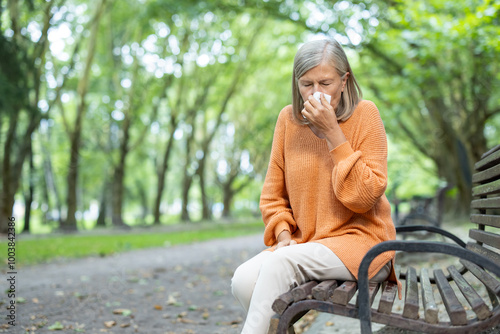 Senior woman in orange sweater sitting on park bench sneezing into tissue. Outdoor setting during autumn signifies allergy or cold symptoms, highlighting seasonal health issues and colder weather.