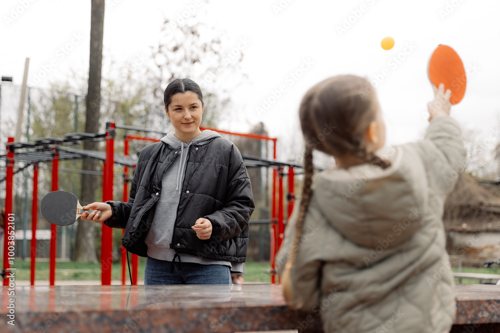 A girl is playing table tennis with a tennis racket and ball in a park, competing with her mother in a game. Joint weekend of mom and daughter A girl is playing table tennis with a tennis racket and ball in a park, competing with her mother in a game. Joint weekend of mom and daughter