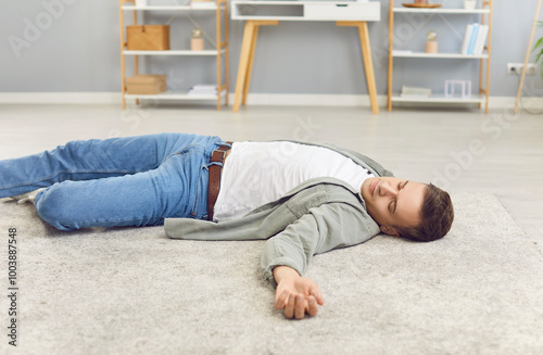 Portrait of a young unconscious man in casual clothes lying alone on floor in living room at home with heart attack or cardiac arrest. Male person fainted indoors. Health and first aid concept.