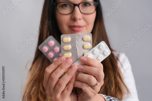 A white woman with glasses holding multiple blister packs of pills in focus, while her face is slightly blurred in the background. The image highlights different colored tablets