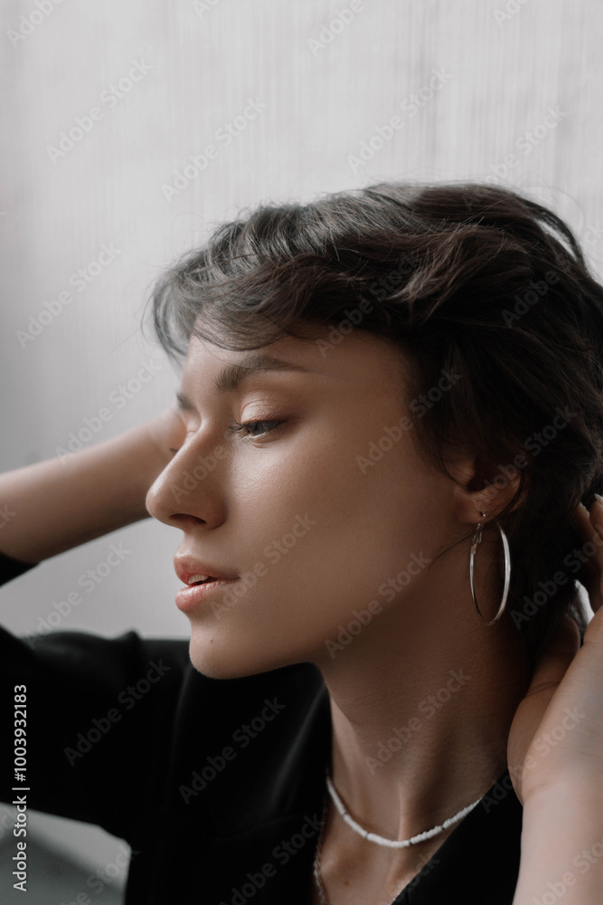 Woman elegantly poses indoors, softly illuminated by natural light. She gently holds her hair back, displaying a serene expression and minimalist jewelry that enhances her beauty Woman elegantly poses indoors, softly illuminated by natural light. She gently holds her hair back, displaying a serene expression and minimalist jewelry that enhances her beauty