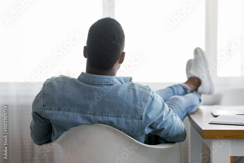 Idle time concept. Rear view of lazy african american employee sitting with his legs on work desk and looking to window, copy space