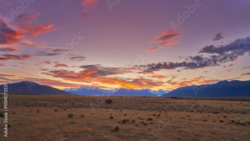 A breathtaking sunset paints the sky above a vast Patagonian landscape, with snow-capped mountains and a solitary group of trees silhouetted against the vibrant colors.