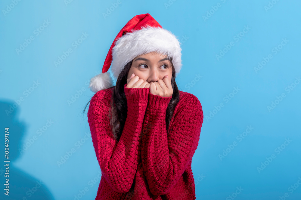 Asian woman dressed in a red knit sweater and wearing a Santa hat smiles sweetly while resting her fists against her cheeks. The background is a solid light blue, creating a cheerful holiday scene Asian woman dressed in a red knit sweater and wearing a Santa hat smiles sweetly while resting her fists against her cheeks. The background is a solid light blue, creating a cheerful holiday scene