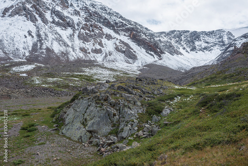 Dramatic cold landscape with rocky hill in green valley against snow covered large mountain range under rainy cloudy gray sky. Gloomy rainy weather in high mountains. Big stone hill in rain time.
