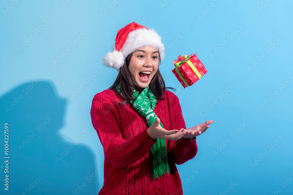 Obraz premium Asian woman in a red sweater, Santa hat, and green scarf is joyfully tossing a red gift box with snowflake patterns and a green ribbon in the air. She stands against a light blue background