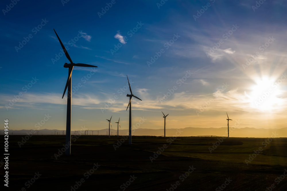 Obraz premium Silhouette Modern windmills with propellers standing in rural farmland agri meadow over orange sunset sky. Wind blowing generating renewable energy for clean resources. Electric power production