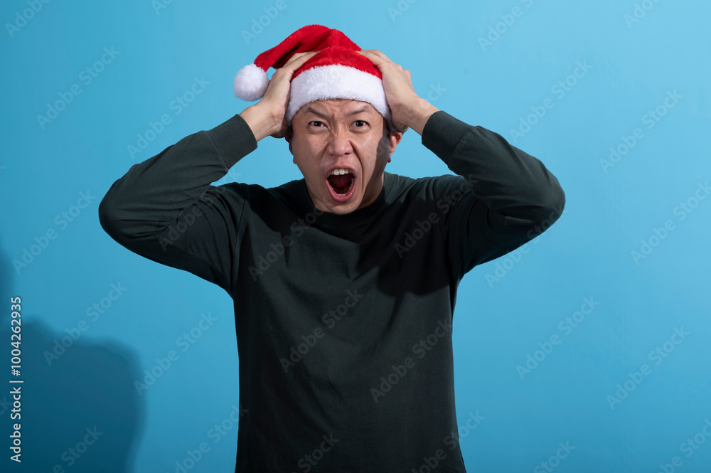 An Asian man wearing a red Santa hat and a dark green long-sleeved shirt reacts with frustration or surprise, pulling his hair with both hands while shouting. He stands against a light blue background