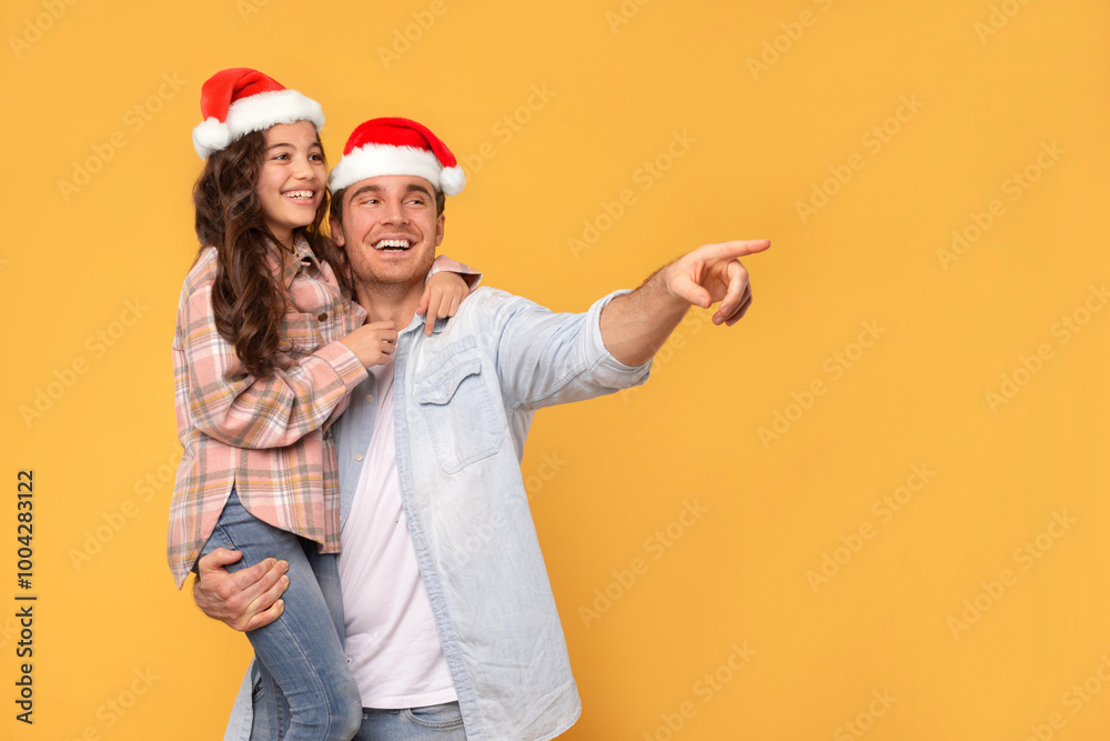 Christmas offer. Happy father holding daughter on hands, both in Santa hats and male pointing, showing free space for Xmas advertisement, yellow background Christmas offer. Happy father holding daughter on hands, both in Santa hats and male pointing, showing free space for Xmas advertisement, yellow background