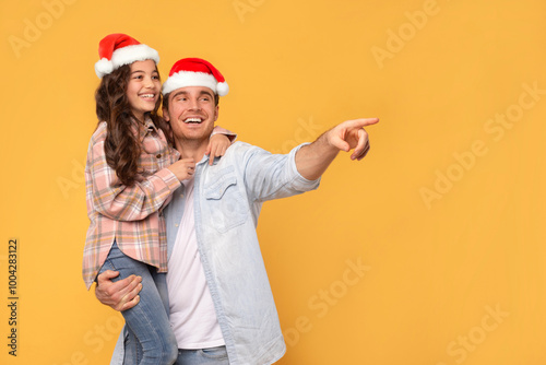 Christmas offer. Happy father holding daughter on hands, both in Santa hats and male pointing, showing free space for Xmas advertisement, yellow background photo