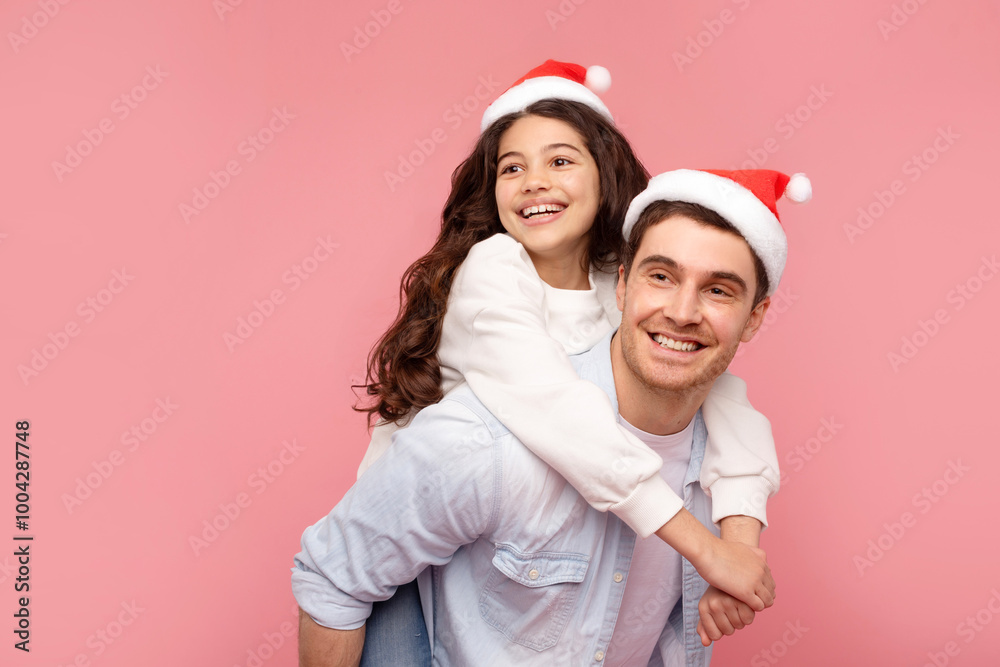 Adorable girl fooling around with her dad, both wearing Santa hats, looking aside at free space on pink background. December, noel time Adorable girl fooling around with her dad, both wearing Santa hats, looking aside at free space on pink background. December, noel time