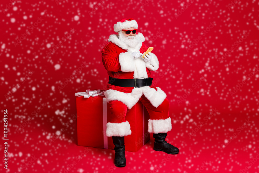 Full length photo of retired old man white beard hold telephone sit large box what chatting elf friend wear x-mas santa costume sunglass headwear leather boot isolated red color background Full length photo of retired old man white beard hold telephone sit large box what chatting elf friend wear x-mas santa costume sunglass headwear leather boot isolated red color background
