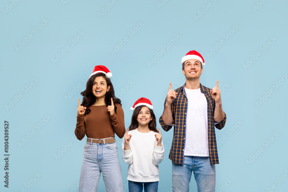 Obraz premium Excited European parents and girl wearing Santa hats and pointing up at copy space above their heads, family demonstrating place for Xmas advertisement, blue background