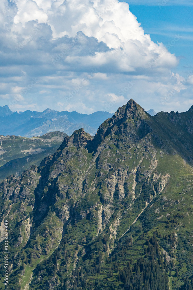 Naklejka premium cross on the peak of rocky mountain with people around, hiker on the summit, hikers paradise, mountain silhouette with mountain range and Austrians alps, cloudy background