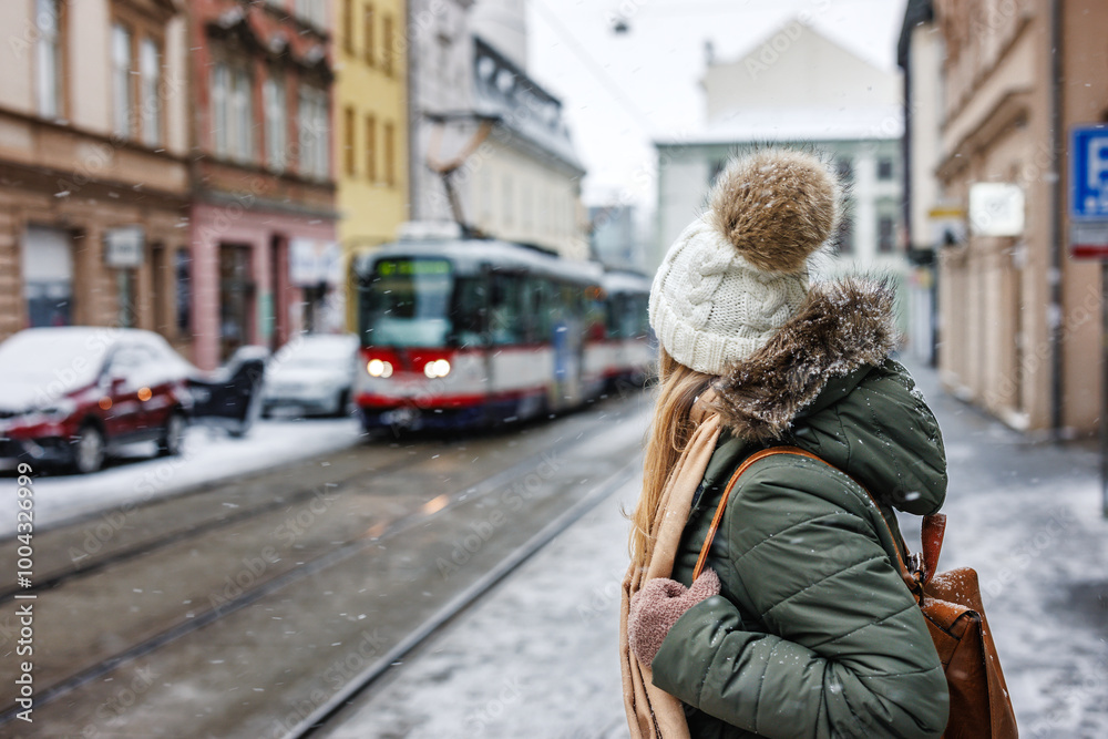 Fototapeta premium Woman looking at passing tram on city street during snowing. Commuter travel by public transportation in winter