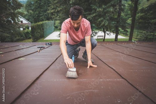 Focused man kneels on a roof while meticulously applying a fresh coat of paint with a wide brush. Surrounded by nature, he demonstrates patience and care, working to enhance the surface photo