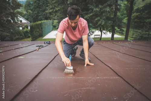 Focused man kneels on a roof while meticulously applying a fresh coat of paint with a wide brush. Surrounded by nature, he demonstrates patience and care, working to enhance the surface photo