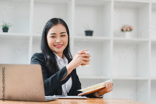 professional woman smiles confidently while holding coffee cup and notebook in modern office setting. bright and organized workspace enhances her positive demeanor.