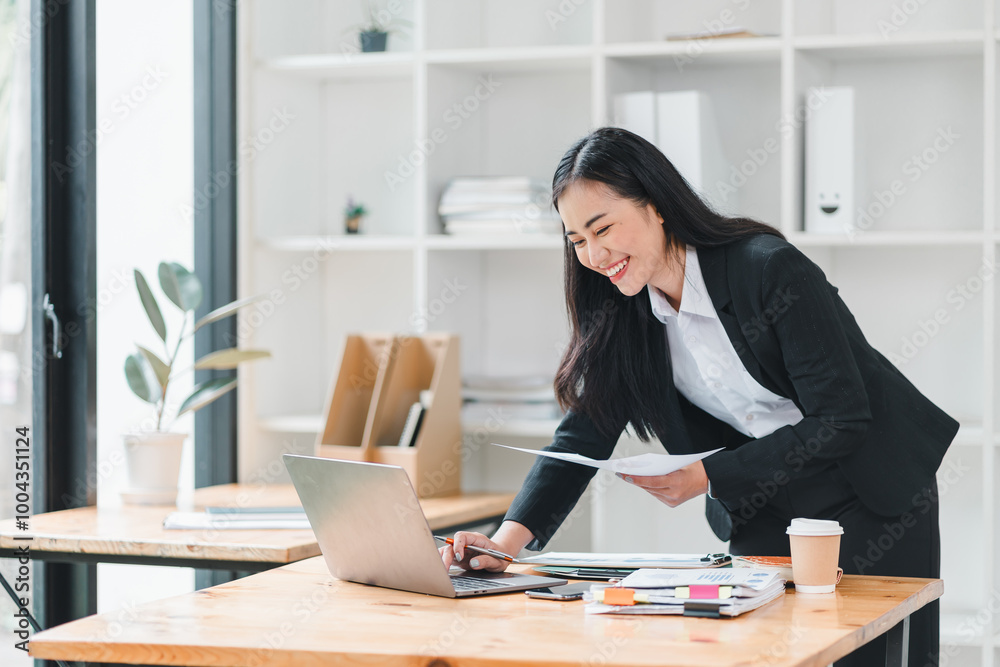 professional woman in suit is working at desk, smiling while using laptop and reviewing documents. modern office setting is bright and organized, creating productive atmosphere. professional woman in suit is working at desk, smiling while using laptop and reviewing documents. modern office setting is bright and organized, creating productive atmosphere.