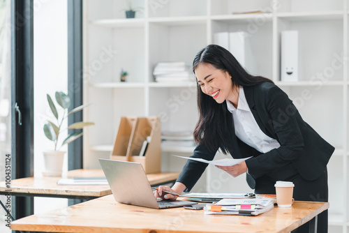 professional woman in suit is working at desk, smiling while using laptop and reviewing documents. modern office setting is bright and organized, creating productive atmosphere.