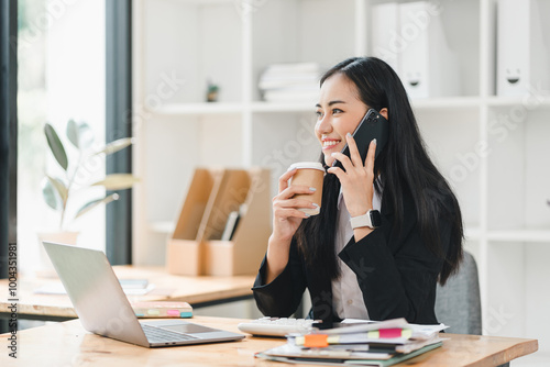 professional woman is engaged in phone conversation while holding coffee cup, surrounded by office supplies and laptop. Her cheerful demeanor reflects productive work environment.