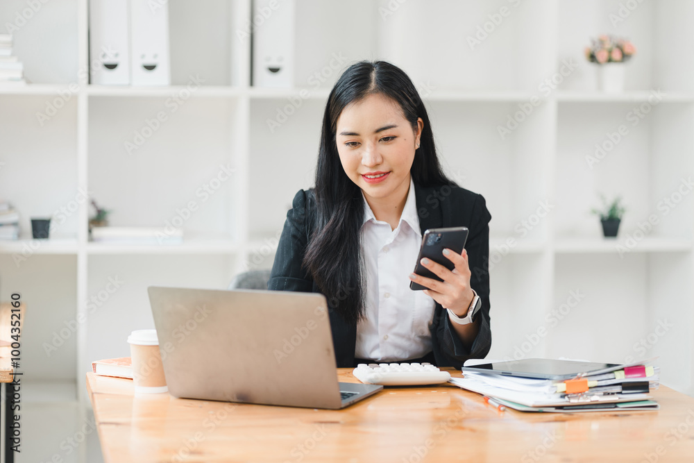 professional woman is engaged in work at her desk, using smartphone while looking at her laptop. scene conveys sense of productivity and focus in modern office environment. professional woman is engaged in work at her desk, using smartphone while looking at her laptop. scene conveys sense of productivity and focus in modern office environment.