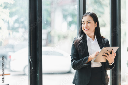 confident businesswoman stands by large window, holding tablet and smiling. She is dressed in professional suit, exuding sense of positivity and determination in modern office environment.