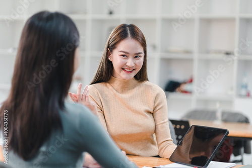 Engaged conversation between two women in modern workspace, showcasing collaboration and teamwork. smiling woman in beige sweater exudes positivity and friendliness.