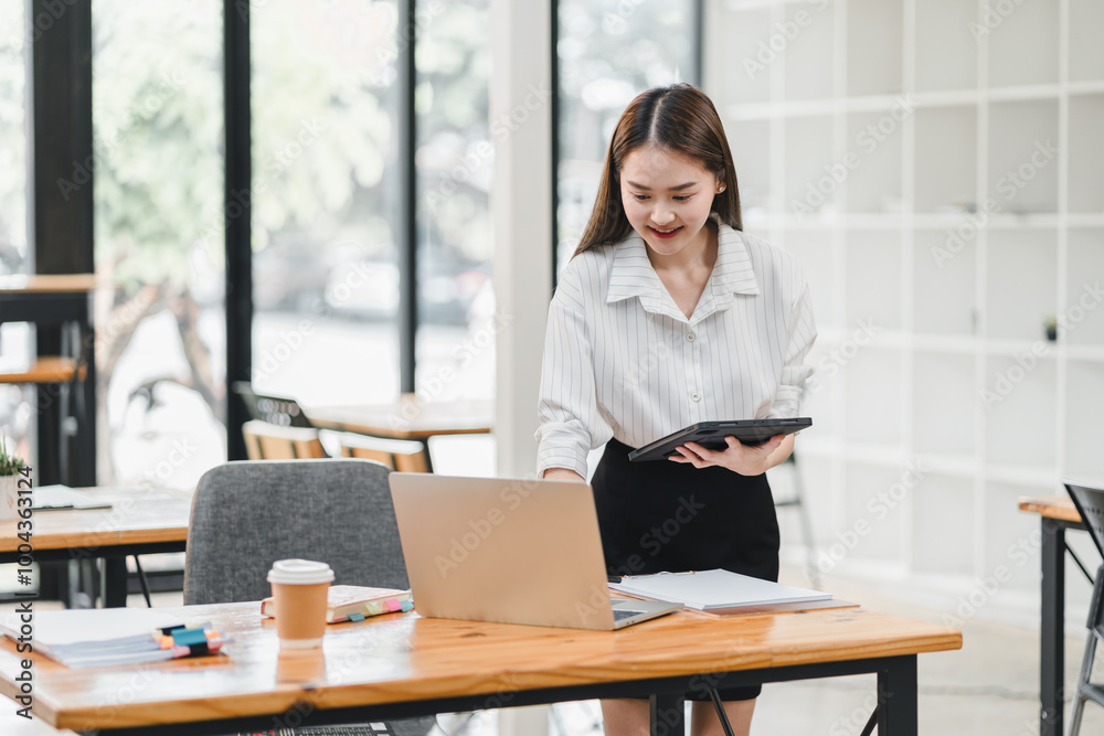 professional woman in modern office setting is working on her laptop while holding tablet. She appears focused and engaged, surrounded by bright and airy workspace. professional woman in modern office setting is working on her laptop while holding tablet. She appears focused and engaged, surrounded by bright and airy workspace.