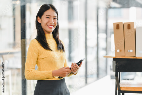 young woman in yellow sweater stands confidently in modern office, holding smartphone and smiling warmly. Her positive demeanor adds vibrant touch to workspace.