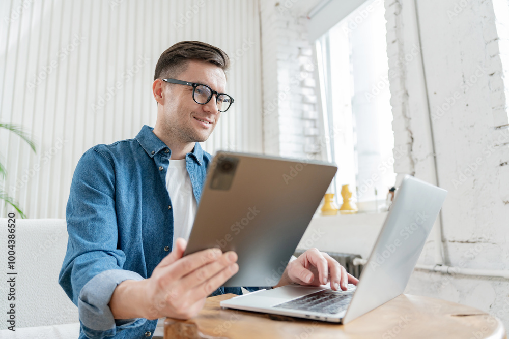 A Man in Casual Attire Uses a Tablet While Working on a Laptop at a Stylish Home Office During Daytime A Man in Casual Attire Uses a Tablet While Working on a Laptop at a Stylish Home Office During Daytime