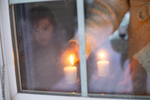 A cozy atmosphere as a person lights candles by a frosty window, with another person's reflection visible.