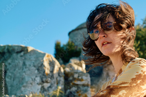 Wind-swept woman with wavy hair and glasses outdoors with ancient stone structures in the background. photo