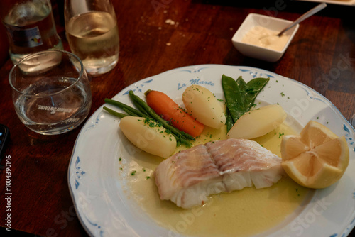 A meal of poached fish with lemon, boiled potatoes, carrots, and spinach served on a white plate with a glass of water and white wine on a wooden table.