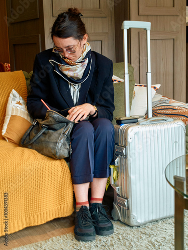 Woman sitting on a yellow couch looking at her phone with a white suitcase and brown bag beside her.