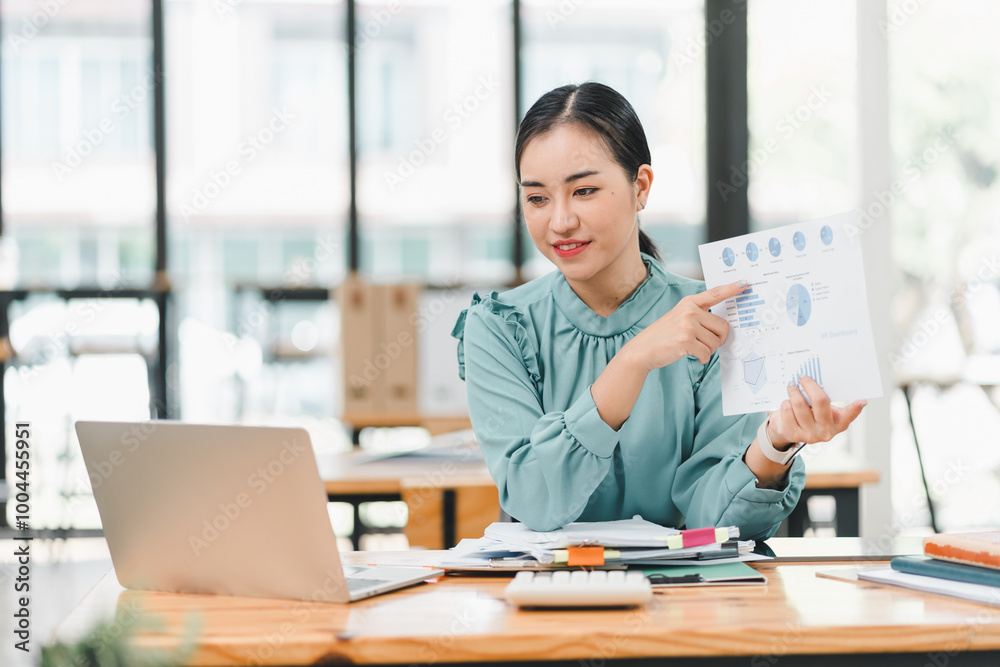 professional woman in light green blouse is presenting data charts while sitting at desk with laptop and documents. She appears engaged and confident, showcasing her analytical skills in modern