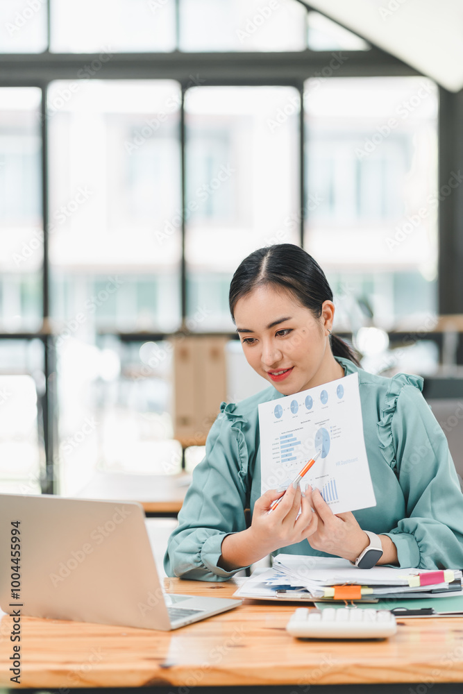 professional woman in green blouse is presenting report with charts while sitting at desk with laptop and documents. She appears engaged and focused on her work. professional woman in green blouse is presenting report with charts while sitting at desk with laptop and documents. She appears engaged and focused on her work.