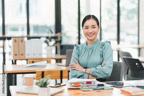 confident woman in green blouse sits at desk in modern office, surrounded by documents and stationery, exuding professionalism and positivity.