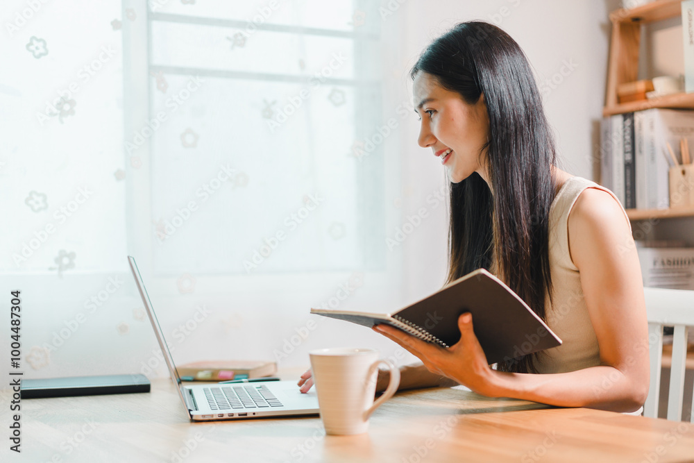 woman is sitting at wooden table, engaged in work on her laptop while holding notebook. She appears focused and content in bright, airy room. woman is sitting at wooden table, engaged in work on her laptop while holding notebook. She appears focused and content in bright, airy room.