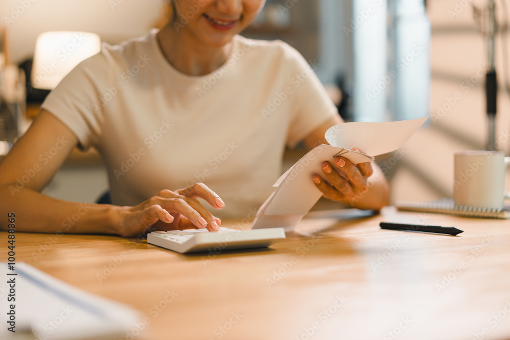 woman is sitting at wooden table, smiling while using calculator and holding documents. warm atmosphere suggests productive and positive environment.