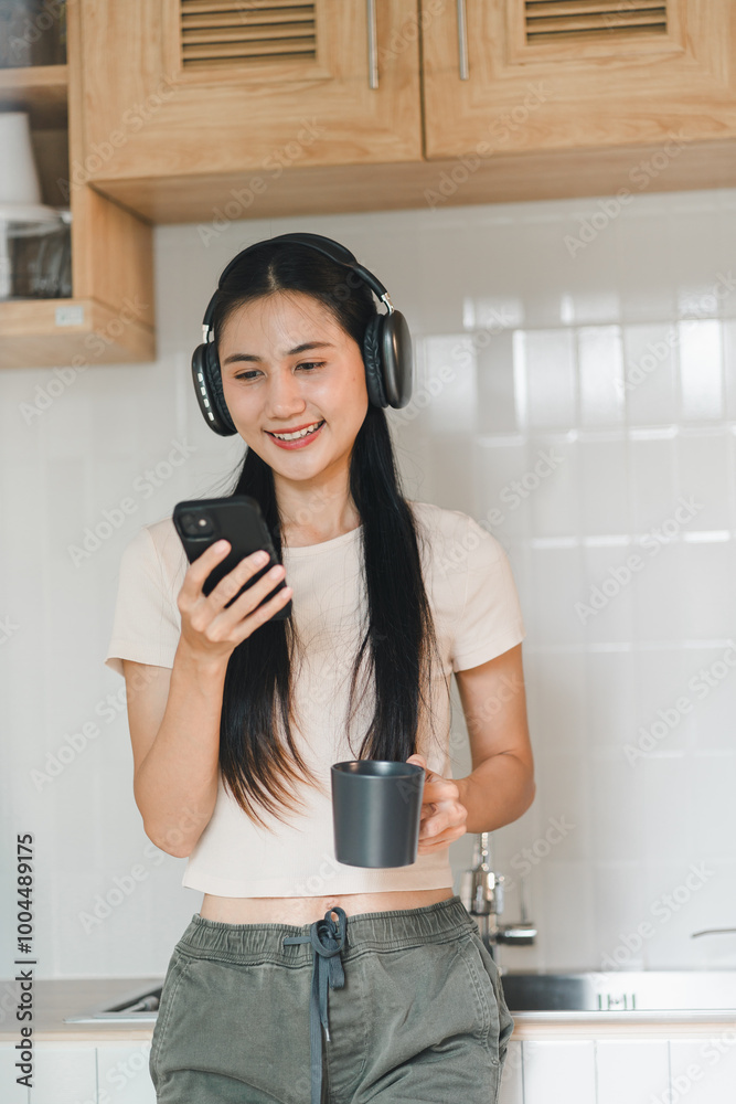 Enjoying moment of relaxation, young woman with long hair wears headphones while checking her phone and holding cup in modern kitchen. Enjoying moment of relaxation, young woman with long hair wears headphones while checking her phone and holding cup in modern kitchen.
