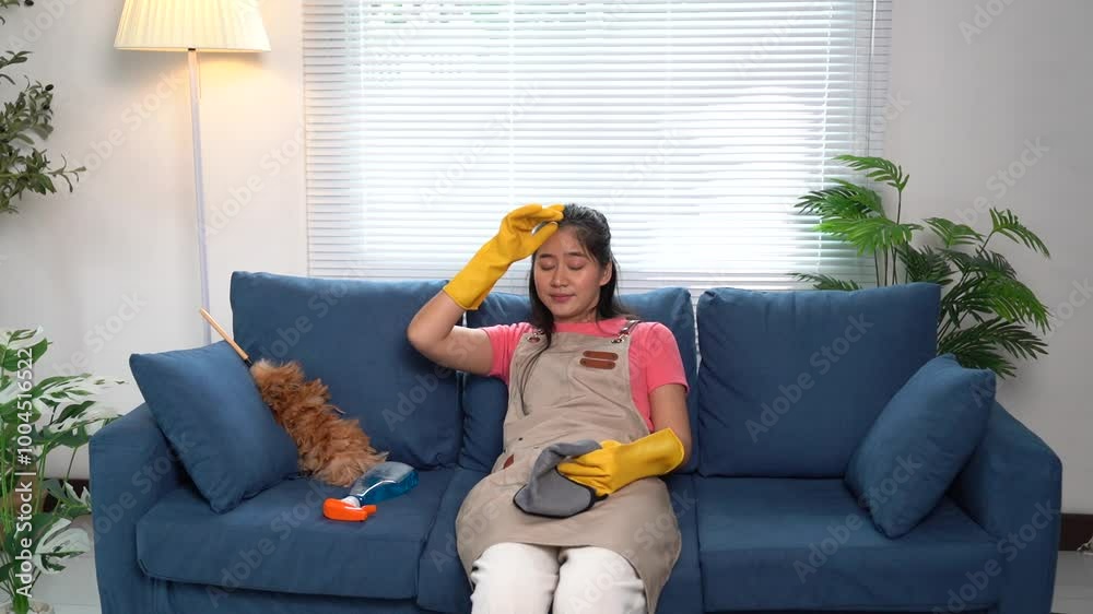 Tired housekeeper relaxing on a sofa touches her forehead, displaying a weary expression. Taking a break from housework, she embodies the exhaustion of domestic chores Tired housekeeper relaxing on a sofa touches her forehead, displaying a weary expression. Taking a break from housework, she embodies the exhaustion of domestic chores