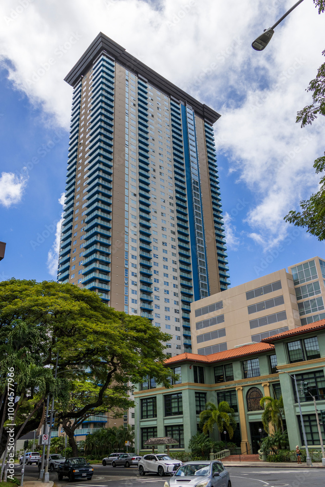 Obraz premium Hotels, condominiums and skyscrapers in the city skyline with lush green trees, cars driving on the street, blue sky and clouds in downtown Honolulu Hawaii USA