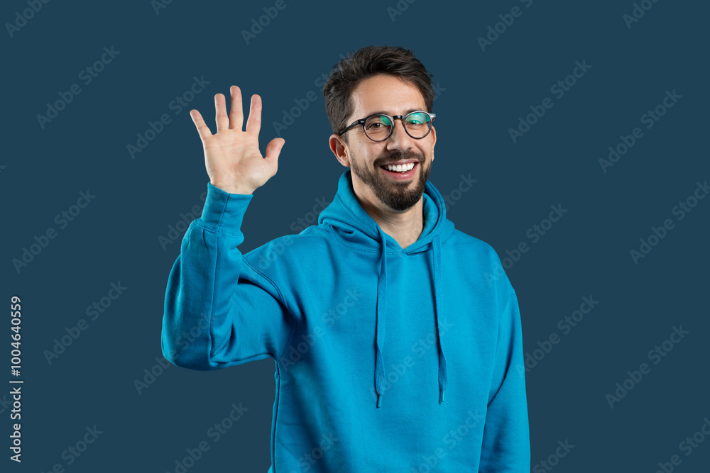 Naklejka premium A cheerful young man greets with a smile and raised hand, dressed in a blue hoodie, set against a dark, solid backdrop. His happy expression conveys friendliness and warmth.